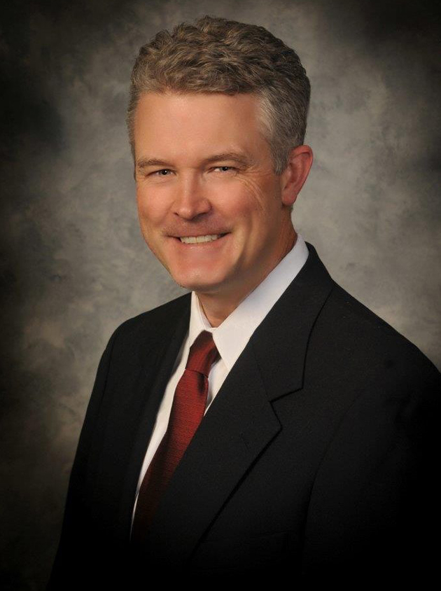 Professional headshot of man in dark suit and red tie against neutral gray background.