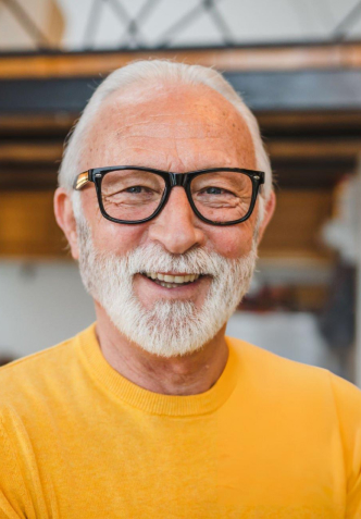 Smiling older man with gray beard and glasses wearing yellow shirt in modern dental clinic interior.