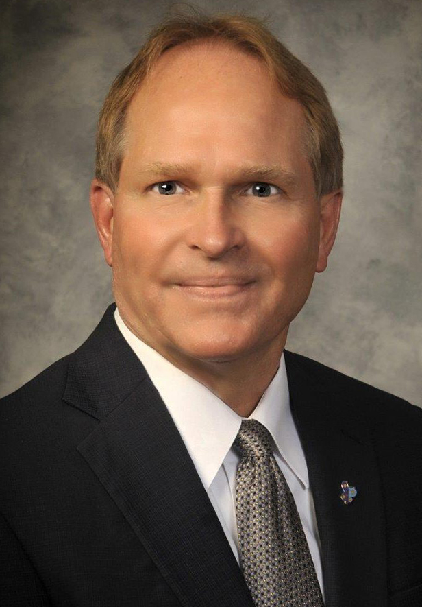 Professional headshot of man in dark suit and tie against neutral gray background.