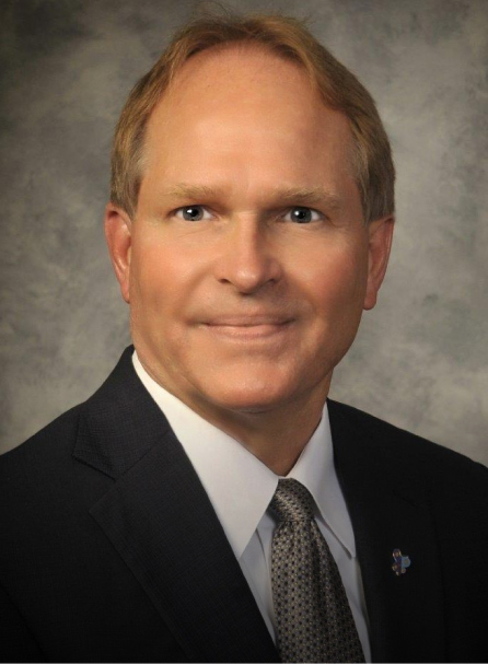 Professional headshot of male dental professional in business suit against neutral gray background.