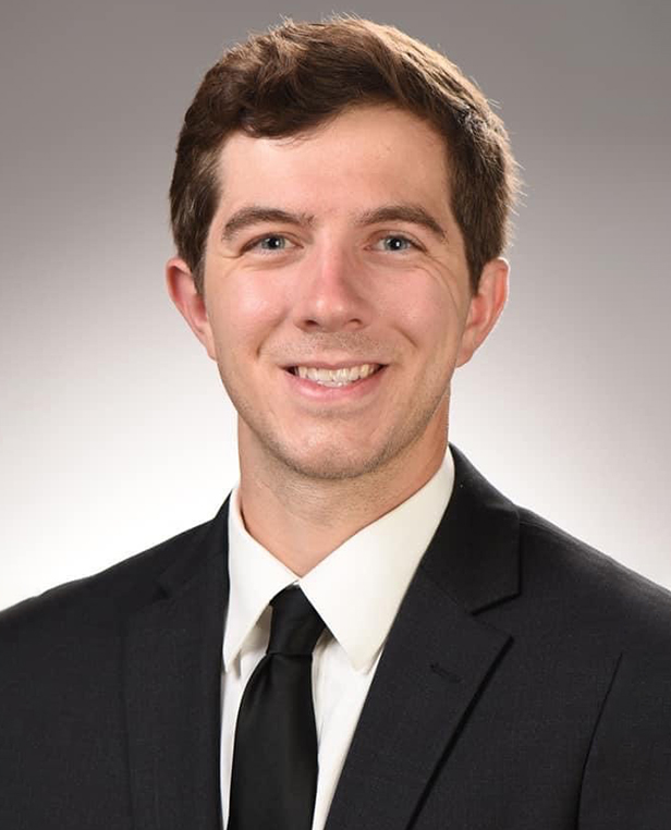 Professional headshot of man in black suit and tie against neutral gray background.