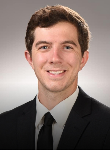 Professional headshot of smiling man in dark suit and tie against gray background.