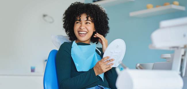Woman holding mirror examining teeth in modern dental clinic office setting.