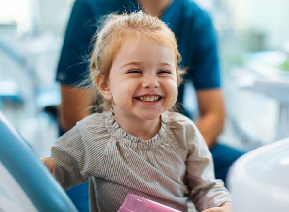 Young girl smiling at dental clinic with dentist in blue scrubs in background.