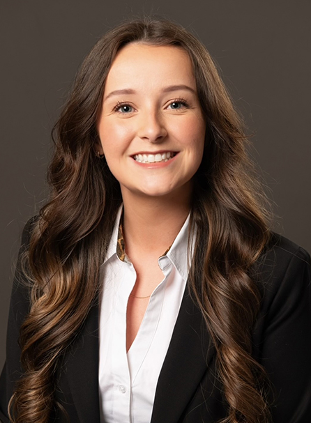 Professional headshot of a woman with long brown hair wearing a black blazer and white shirt against a gray background.